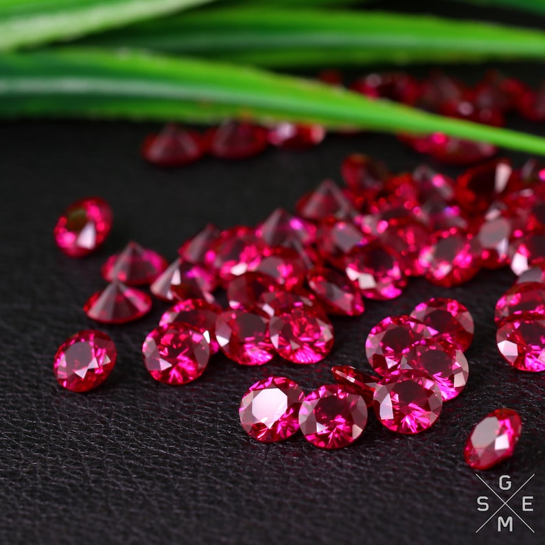 May include: A collection of bright red round gemstones, likely rubies, scattered on a black surface. The gemstones are faceted and reflect light.