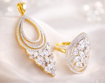May include: A gold pendant and ring set. The pendant has a teardrop shape with three inner loops and a cluster of white gemstones at the bottom. The ring features a similar design, with a large oval face covered in white gemstones.