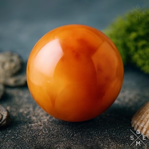 Può includere: Una gemma sferica lucida color ambra. L'orbita ha una superficie lucida, che riflette la luce. La gemma è posta su una superficie strutturata grigio scuro con accenni di muschio verde e piccole pietre.