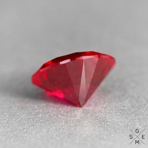 May include: A deep red ruby gemstone with a faceted cut. The gemstone is lying on a grey surface.