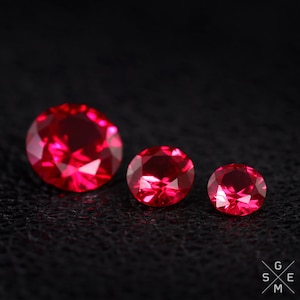 May include: Three red ruby gemstones of different sizes, arranged in a row on a black surface.