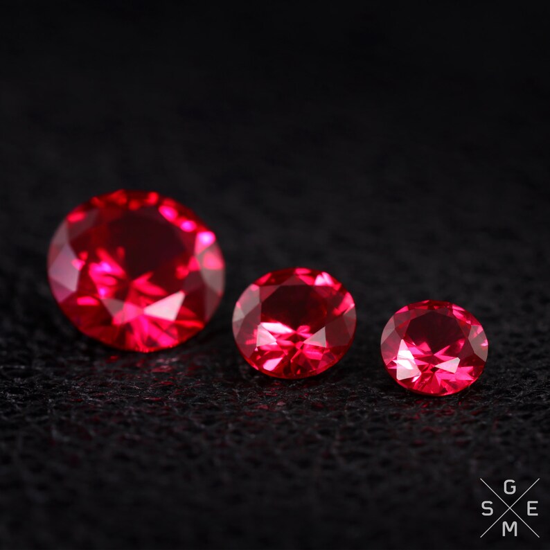 May include: Three red ruby gemstones of different sizes, arranged in a row on a black surface.