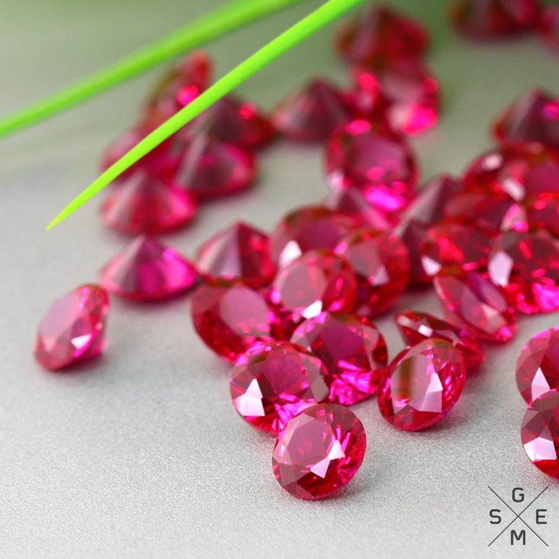 May include: A close-up of a group of bright red gemstones, likely rubies, scattered on a grey surface. The gemstones are round and have a polished, faceted appearance.