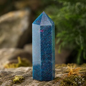 Op de afbeelding: Een gepolijste zeshoekige kristaltoren met een diepblauwe basiskleur en gespikkelde donkerblauwe en magentakleurige insluitingen door de hele steen. De toren heeft een puntige bovenkant en een glad, reflecterend oppervlak.