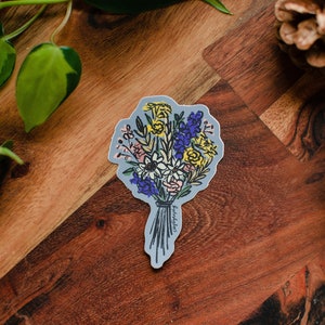 A semi-abstract drawing of a bouquet of flowers as a sticker sits on a piece of wood. The sticker has a periwinkle blue background and is die cut in the shape of the bouquet design. Surrounding the sticker are a few pinecones and a pothos vine.