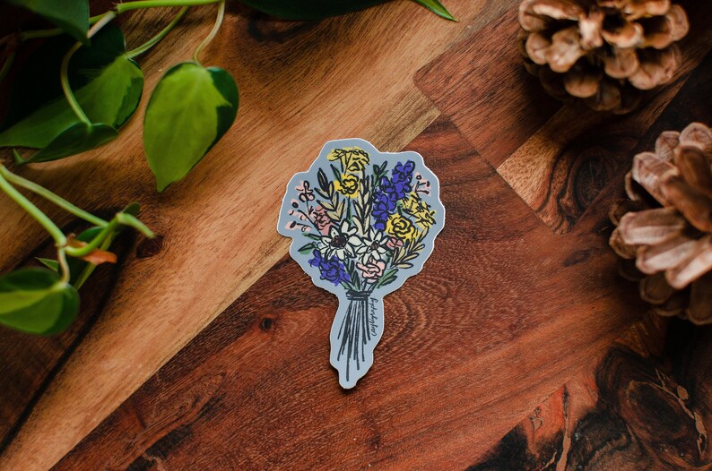 A semi-abstract drawing of a bouquet of flowers as a sticker sits on a piece of wood. The sticker has a periwinkle blue background and is die cut in the shape of the bouquet design. Surrounding the sticker are a few pinecones and a pothos vine.