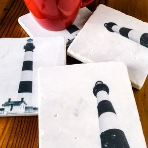 May include: Four white square coasters with a black and white lighthouse image printed on each. The coasters are on a wooden surface.