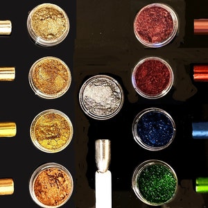 Op de afbeelding: Assortiment van kleine potjes gevuld met kleurrijk glitterpoeder. De potjes zijn in rijen op een zwart oppervlak gerangschikt. Er zijn ook verschillende kleine metalen gereedschappen, sommige met een metalen afwerking en andere met een gekleurde afwerking.