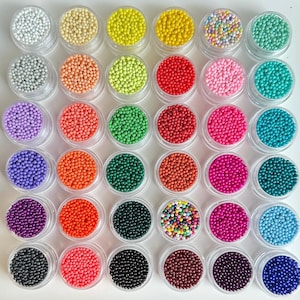 Pode incluir: Uma vista superior de 36 pequenos potes de plástico transparente cheios de pequenas contas redondas coloridas. As contas são de várias cores, incluindo branco, amarelo, vermelho, rosa, verde e azul. Os potes estão dispostos em um padrão de grade.