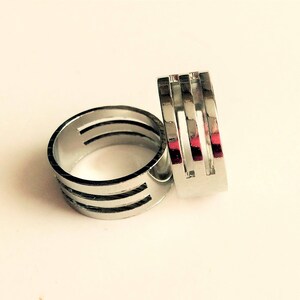 May include: Two silver rings with a modern design. One ring is round with three horizontal cutouts, and the other is rectangular with three vertical cutouts. The rings are displayed on a white surface.