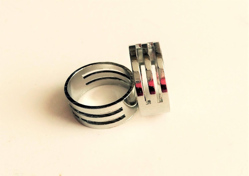 May include: Two silver rings with a modern design. One ring is round with three horizontal cutouts, and the other is rectangular with three vertical cutouts. The rings are displayed on a white surface.