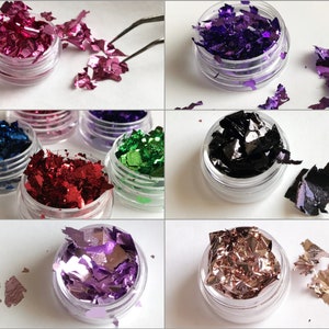 May include: Assortment of small jars filled with colorful metallic flakes. The flakes are in various shades of pink, purple, green, red, black, and gold. The jars are clear plastic with a screw-on lid.
