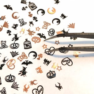 May include: A variety of black and gold Halloween-themed nail art embellishments, including bats, spiders, pumpkins, and hearts, are scattered on a white surface. A pair of tweezers is used to pick up the embellishments.