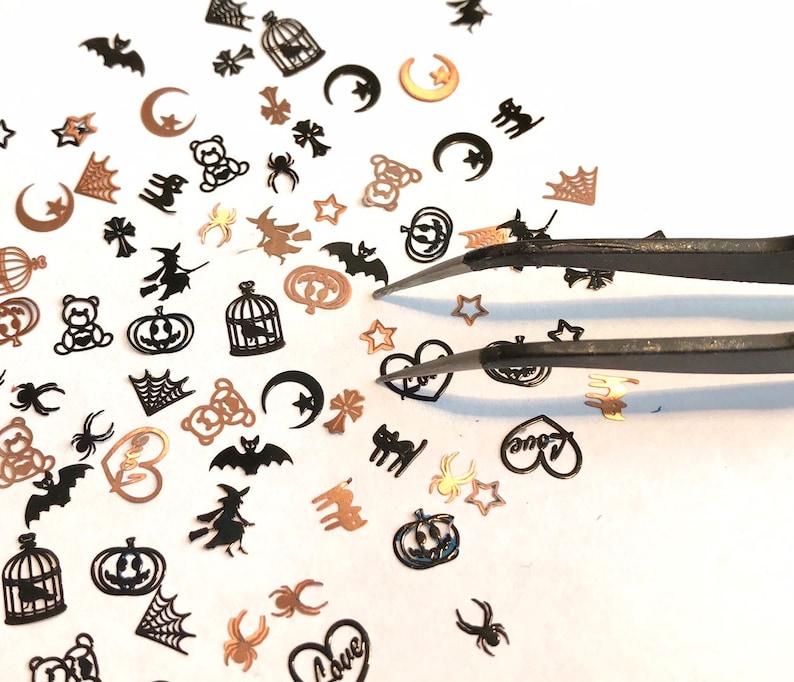 May include: A variety of black and gold Halloween-themed nail art embellishments, including bats, spiders, pumpkins, and hearts, are scattered on a white surface. A pair of tweezers is used to pick up the embellishments.