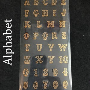 May include: A sheet of gold alphabet and number stickers on a black background. The letters and numbers are in a decorative font.