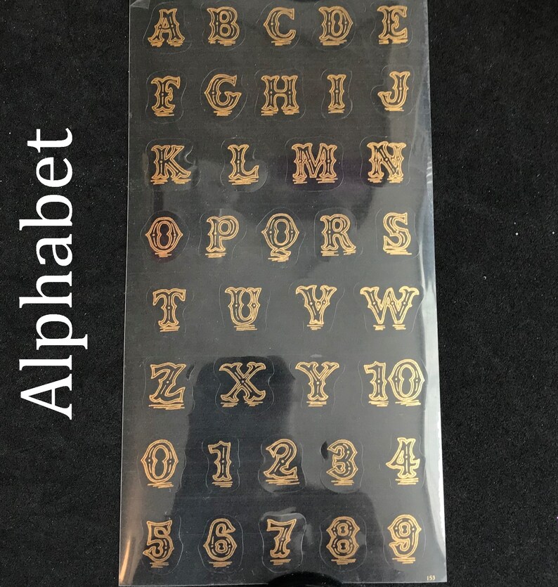 May include: A sheet of gold alphabet and number stickers on a black background. The letters and numbers are in a decorative font.