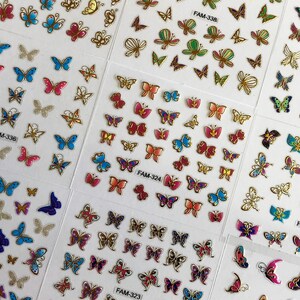 May include: A sheet of colorful butterfly nail stickers with different designs and colors. The stickers are arranged in rows and columns on a white background. The sheet includes the text "FAM-336", "FAM-328", "FAM-323", "FAM-324", "FAM-338", "FAM-319", and "FAM".