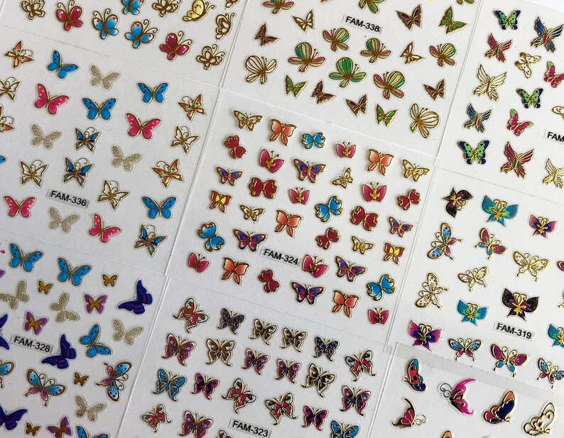 May include: A sheet of colorful butterfly nail stickers with different designs and colors. The stickers are arranged in rows and columns on a white background. The sheet includes the text "FAM-336", "FAM-328", "FAM-323", "FAM-324", "FAM-338", "FAM-319", and "FAM".