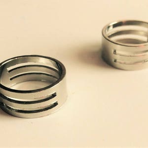 May include: Two silver rings with a modern design. Each ring has three horizontal cut-out lines, creating a unique, open pattern. The rings are displayed on a light-colored surface, showcasing their metallic shine.