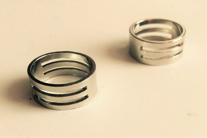 May include: Two silver rings with a modern design. Each ring has three horizontal cut-out lines, creating a unique, open pattern. The rings are displayed on a light-colored surface, showcasing their metallic shine.