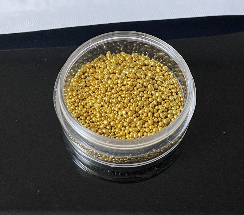 Op de afbeelding: Een doorzichtige plastic container gevuld met kleine, ronde, gouden kralen.