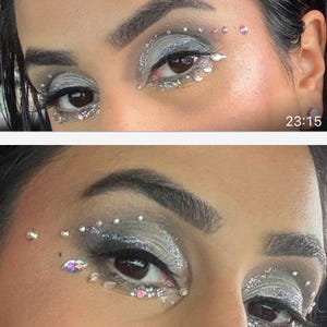 May include: Close-up of two eyes with silver and gray eyeshadow, glitter, and rhinestones. The makeup design includes rhinestones above and below the eyes, with a time stamp of 23:15 in the bottom right corner.