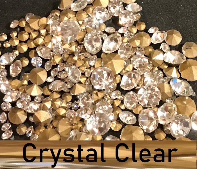 May include: A close-up image of a mix of clear and gold rhinestones. The rhinestones are scattered on a black surface. The text "Crystal Clear" is visible at the bottom of the image.