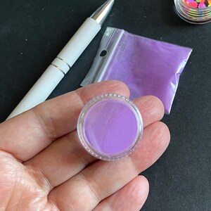 May include: A small clear jar filled with purple glitter. The jar is sitting on a black surface.