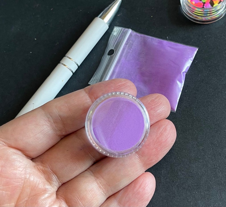 May include: A small clear jar filled with purple glitter. The jar is sitting on a black surface.