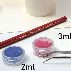 May include: Two small jars of colourful powder, one blue and one pink, with labels indicating 2 ml and 3 ml. A red pencil and tweezers are also visible.