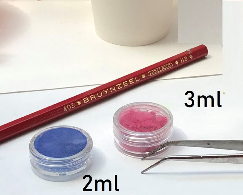 May include: Two small jars of colourful powder, one blue and one pink, with labels indicating 2 ml and 3 ml. A red pencil and tweezers are also visible.