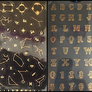 May include: Two sheets of gold sticker paper. One sheet features constellations and celestial symbols. The other sheet features the alphabet and numbers in a decorative font.