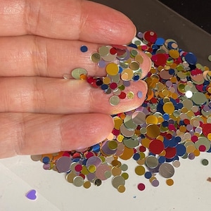 Op de afbeelding: Een verscheidenheid aan kleurrijke, iriserende ronde glitter in goud, zilver, rood, blauw en paars. De glitter is verspreid over een wit oppervlak.
