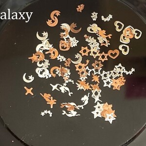 May include: A variety of small, metallic charms in silver and gold tones. The charms include stars, moons, hearts, and other abstract shapes. The charms are arranged on a black surface. The text "Galaxy" is written in white at the top of the image.