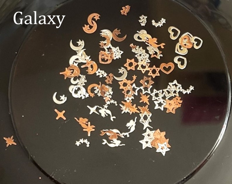 May include: A variety of small, metallic charms in silver and gold tones. The charms include stars, moons, hearts, and other abstract shapes. The charms are arranged on a black surface. The text "Galaxy" is written in white at the top of the image.