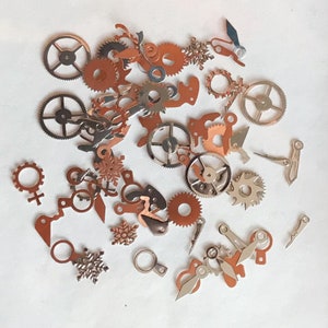 May include: A collection of assorted metal gears and watch parts in silver and copper tones. The parts are scattered on a white surface.