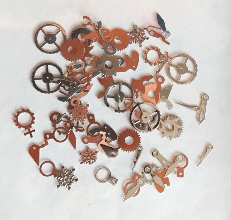 May include: A collection of assorted metal gears and watch parts in silver and copper tones. The parts are scattered on a white surface.