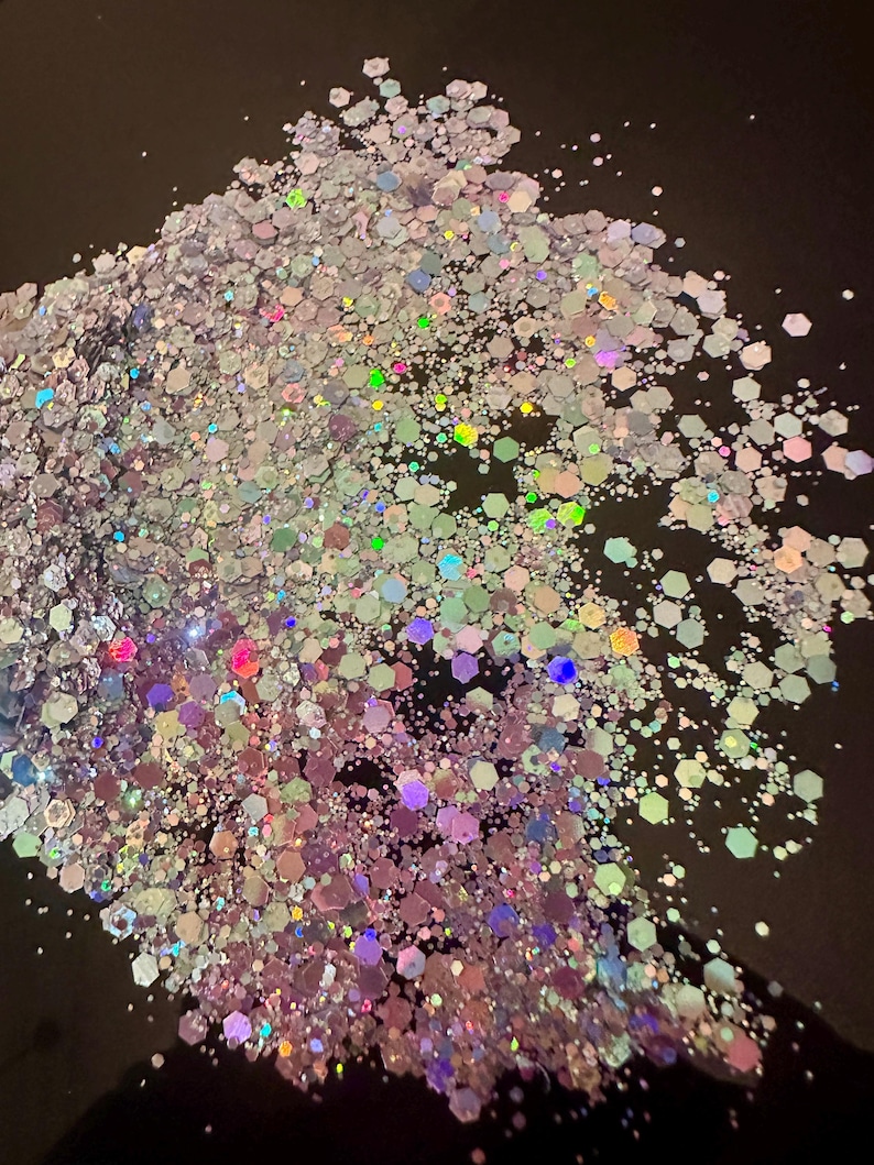 May include: A pile of iridescent glitter with a mix of hexagonal and smaller shapes. The glitter reflects a range of colors, including pink, green, and blue, against a dark background. Ideal for crafts and decorations.