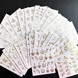 May include: A set of 20 gold temporary tattoo sheets with various designs, including butterflies, flowers, geometric shapes, and a chandelier. The sheets are white with gold designs.