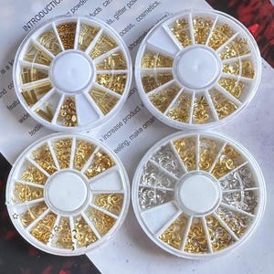Puede incluir: Cuatro ruedas de plástico blanco con 12 compartimentos cada una, llenas de adornos de arte para uñas de oro y plata. Las ruedas están etiquetadas con las palabras "increase product" y "eeling, make orman".