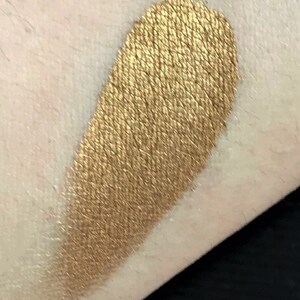 K&ouml;nnte beinhalten: Ein goldener Lidschatten-Swatch auf der Haut. Der Lidschatten hat ein metallisches Finish und ein strukturiertes Aussehen, das Licht reflektiert. Die Farbe ist ein warmes, sattes Gold.