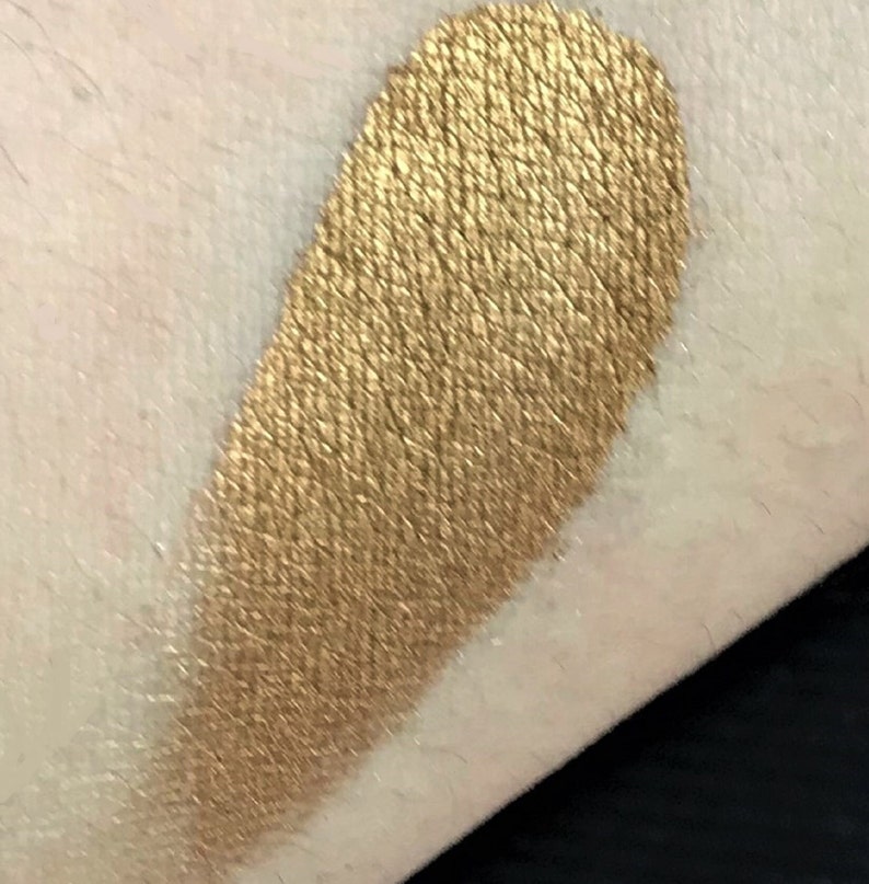 K&ouml;nnte beinhalten: Ein goldener Lidschatten-Swatch auf der Haut. Der Lidschatten hat ein metallisches Finish und ein strukturiertes Aussehen, das Licht reflektiert. Die Farbe ist ein warmes, sattes Gold.