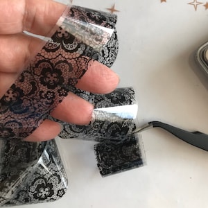 Op de afbeelding: Een rol transparante nagelkunst-transferfolie met een zwart kantpatroon. De folie is opgerold en wordt in een hand gehouden.