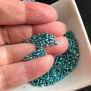 Op de afbeelding: Een close-up van een kleine witte kom gevuld met teal blauwe metallic sprinkles. De sprinkles zijn klein en onregelmatig van vorm.