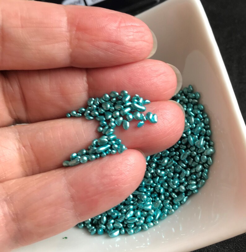 Op de afbeelding: Een close-up van een kleine witte kom gevuld met teal blauwe metallic sprinkles. De sprinkles zijn klein en onregelmatig van vorm.