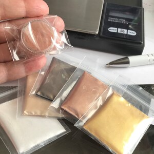 May include: Five small plastic bags containing different coloured metallic powders. The powders are in shades of white, beige, grey, copper, and gold. A small container of a similar powder is being held in a hand.