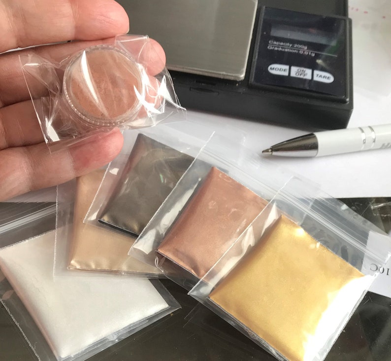 May include: Five small plastic bags containing different coloured metallic powders. The powders are in shades of white, beige, grey, copper, and gold. A small container of a similar powder is being held in a hand.