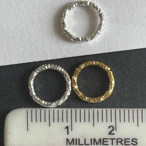 May include: Three silver and gold textured metal rings, measuring approximately 1 centimeter in diameter, are displayed on a black surface with a white ruler.