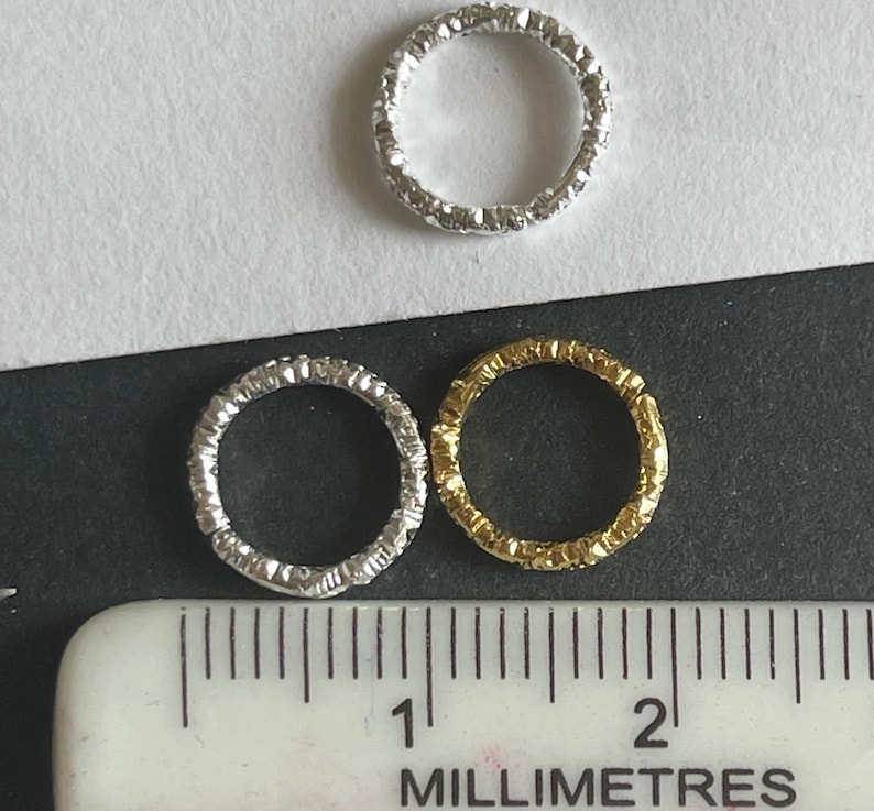 May include: Three silver and gold textured metal rings, measuring approximately 1 centimeter in diameter, are displayed on a black surface with a white ruler.