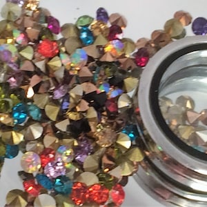 May include: A silver locket charm with a clear glass window filled with colorful rhinestones. The locket is surrounded by a scattering of loose rhinestones in various colors, including gold, red, green, blue, and purple.